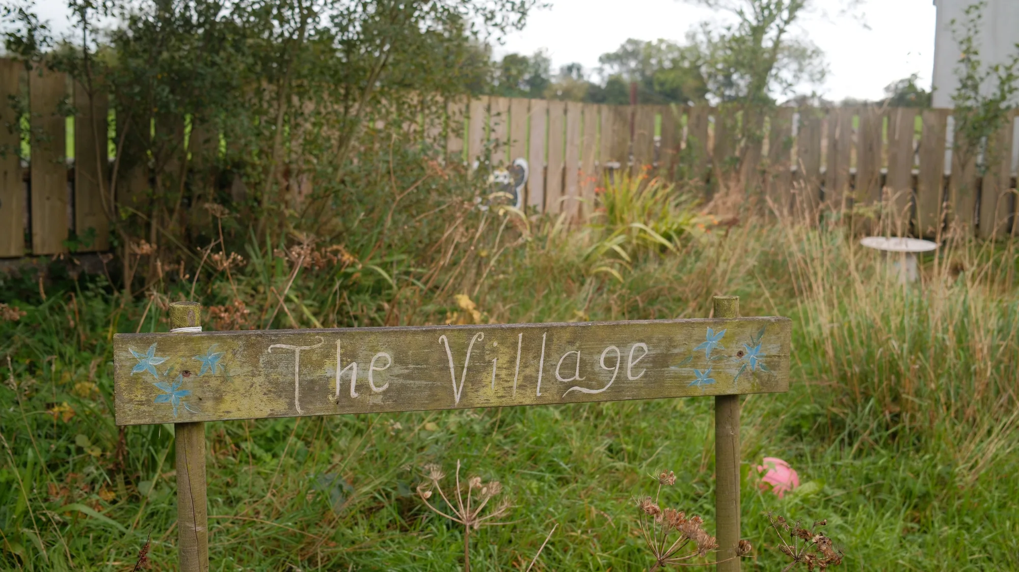 The Village sign among plants by the fence